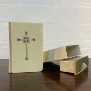 May include: A small, white book with a gold and blue cross design on the cover. The book is in a white box with a gold trim.