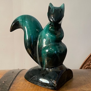 Puede incluir: Una figurita de ardilla de cerámica verde y negra esmaltada. La ardilla está sentada sobre una base y tiene una cola larga y rizada.