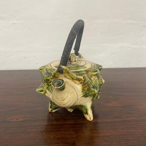 May include: A ceramic teapot with a unique design. The teapot is primarily beige with green and brown accents. It has a textured surface, a black woven handle, and three small feet. The spout is cylindrical.