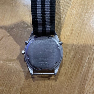 Pu&ograve; includere: Un orologio da polso argentato con un cinturino in nylon a righe nere e grigie. Il quadrante dell'orologio non &egrave; visibile, ma sul retro della cassa dell'orologio &egrave; presente il testo "WATER RESISTANT STAINLESS STEEL JAPAN MOV'T JAPAN T-N21764".