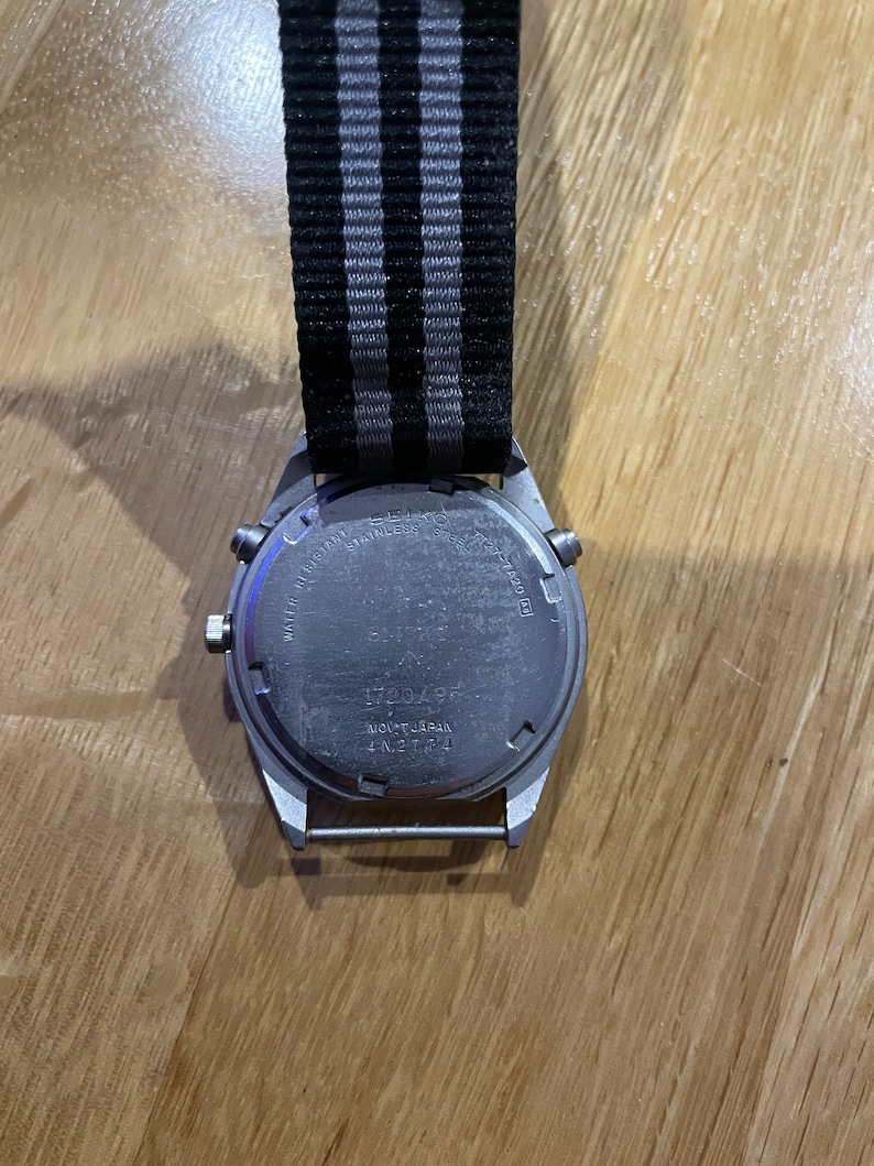 Pu&ograve; includere: Un orologio da polso argentato con un cinturino in nylon a righe nere e grigie. Il quadrante dell'orologio non &egrave; visibile, ma sul retro della cassa dell'orologio &egrave; presente il testo "WATER RESISTANT STAINLESS STEEL JAPAN MOV'T JAPAN T-N21764".