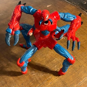 K&ouml;nnte beinhalten: Eine rote und blaue Actionfigur von Spider-Man mit mehreren Beinen und Krallen. Die Figur ist in einer bedrohlichen Pose mit offenem Mund und sichtbaren Z&auml;hnen dargestellt.