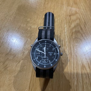 Pu&ograve; includere: Un orologio da polso cronografo nero con cassa argentata e cinturino in nylon a righe nere e grigie. Il quadrante dell'orologio ha delle marcature bianche e una lancetta dei secondi argentata.