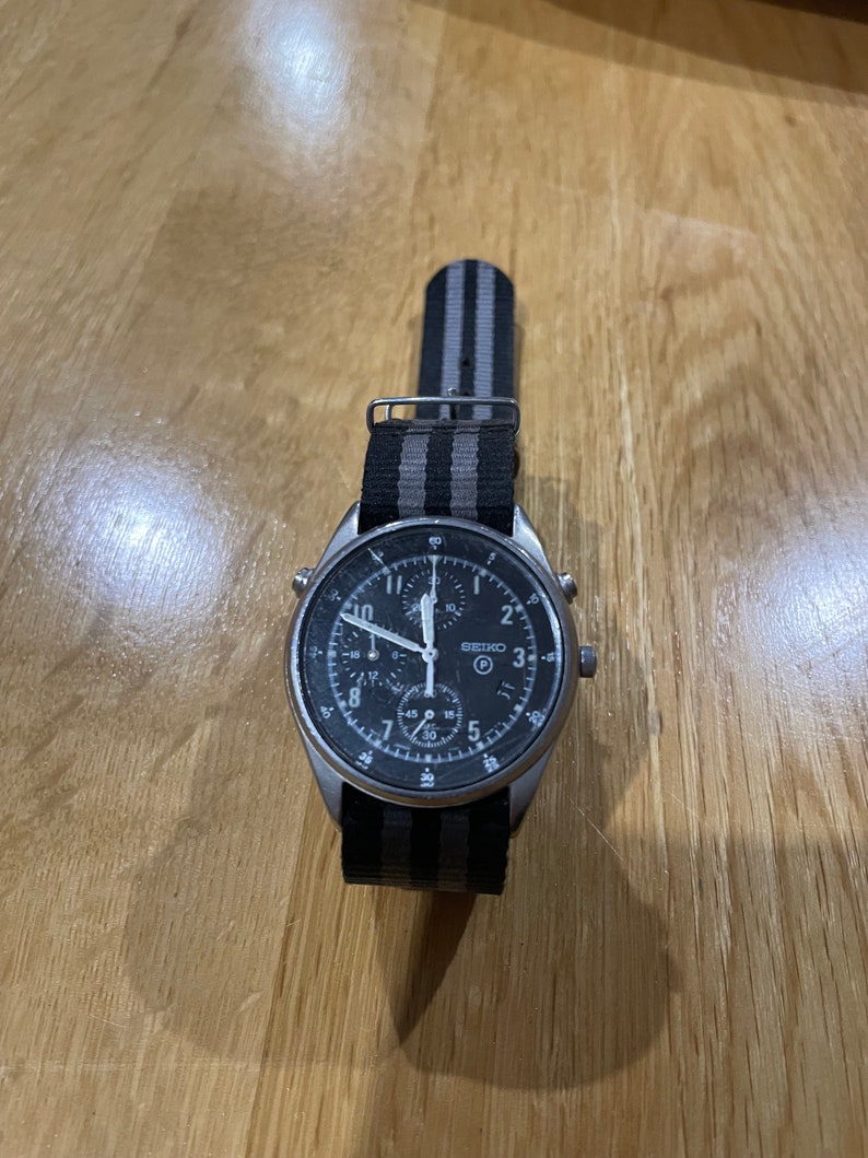 Pu&ograve; includere: Un orologio da polso cronografo nero con cassa argentata e cinturino in nylon a righe nere e grigie. Il quadrante dell'orologio ha delle marcature bianche e una lancetta dei secondi argentata.