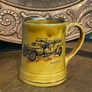Puede incluir: Una taza de cerámica marrón con un borde dorado y una imagen en blanco y negro de un coche Rolls-Royce vintage. La taza tiene el texto "Rolls-Royce" impreso en la parte inferior.