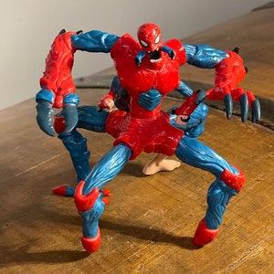 K&ouml;nnte beinhalten: Eine rote und blaue Actionfigur von Spider-Man mit zus&auml;tzlichen Gliedma&szlig;en und einem bedrohlichen Gesichtsausdruck. Die Figur ist in einer bedrohlichen Pose mit ausgestreckten Krallen dargestellt.
