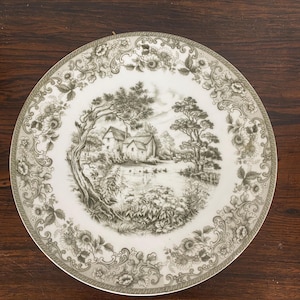 Könnte beinhalten: Ein weißer Keramikteller mit grünem und weißem Blumenrand und einem zentralen Bild eines Häuschens in einer bewaldeten Landschaft.