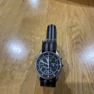 Pu&ograve; includere: Un orologio da polso cronografo argentato con quadrante nero e cinturino in nylon a righe nere e grigie.