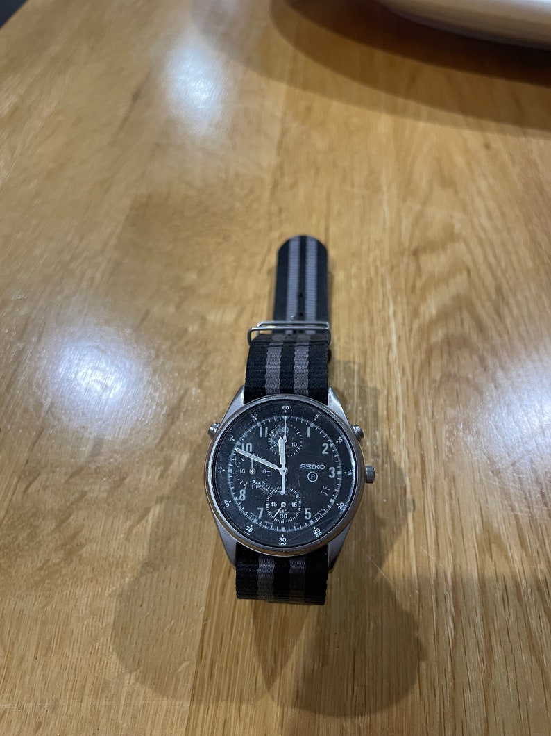 Pu&ograve; includere: Un orologio da polso cronografo argentato con quadrante nero e cinturino in nylon a righe nere e grigie.