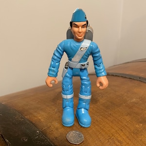 May include: A blue action figure of a man in a uniform with a jetpack. The figure is wearing a blue cap, boots, and a white sash. A coin is on the wooden surface in front of the figure.