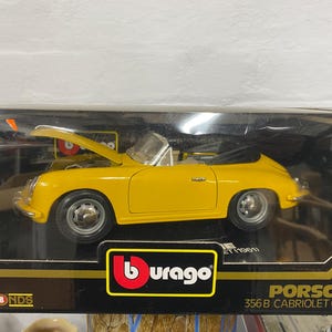 May include: A yellow 1/18 scale diecast model of a Porsche 356 B Cabriolet (1961) in a display box. The car has a black convertible top and black wheels. The box has a Burago logo and the text "Porsche 356 B Cabriolet (1961)".