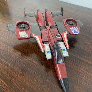 May include: A red and black toy jet fighter with a cockpit and two engines. The jet has a silver stripe on the side and a blue Autobot symbol on the wing.