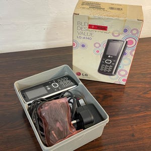May include: An LG-A140 mobile phone with a black and silver design, presented with its original box. The box features the text "Blending of Design and Value" and a graphic of the phone. The phone and accessories are in a grey plastic container.