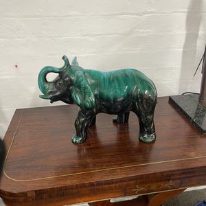 May include: A green and black glazed ceramic elephant figurine. The elephant has its trunk raised and is standing on a dark wood table.