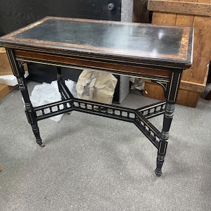 May include: An antique rectangular table with a black leather top and ornate wooden legs. The table features a dark wood frame with gold accents and decorative cross supports. The legs have small caster wheels. A classic piece of furniture, approx. 120cm long.