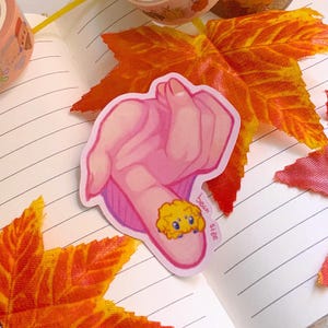 May include: A pink hand sticker with a small yellow bear on the finger. The sticker has the text "bean size" written on it.