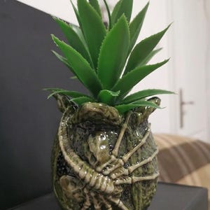 May include: A decorative green ceramic pot with a detailed alien design, holding a vibrant green artificial plant. The pot features a textured surface and a skeletal creature wrapped around it, creating a unique and thematic piece.