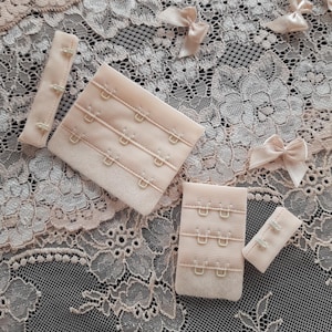 Puede incluir: Cuatro extensores de tirantes de sujetador beige con cierres de gancho y ojo. Los extensores están sobre un fondo de encaje blanco.