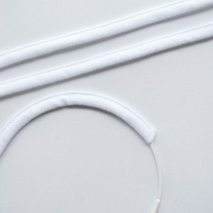 May include: A close-up shot of white, plush headbands and elastic cords against a white background. The headbands are curved and straight, with a soft, velvety texture. The elastic cords are thin and white.