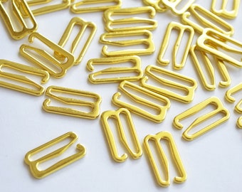 May include: A pile of gold-coloured metal bra strap adjusters. The adjusters are small and have a rectangular shape with two loops on each end.