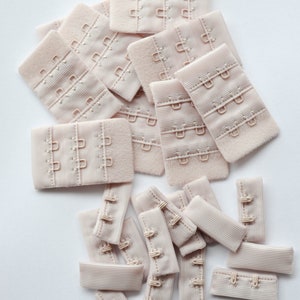 May include: A pile of beige bra strap extenders with hooks and eyes. The extenders are made of a soft, stretchy fabric.