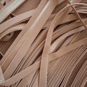 May include: A large collection of nude-colored elastic bands arranged in a pile. Each strip features a textured surface on one side and a smooth, slightly reflective finish on the other, with visible stitching running along the edges of the material.