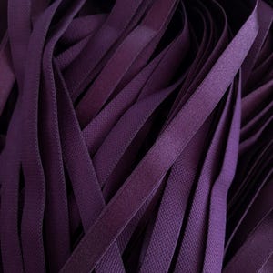 May include: Close-up of a pile of dark purple elastic ribbon. The ribbon is flat, with a slightly textured surface, and appears to be made of a woven material. The ribbons are layered and overlapping, creating a dense, textured pattern.