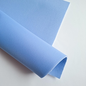 Può includere: Un tessuto di feltro blu chiaro, parzialmente arrotolato, su sfondo bianco. Il feltro ha una consistenza morbida e opaca e un colore uniforme. La parte arrotolata rivela lo spessore e il bordo del tessuto.