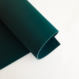 Puede incluir: Una hoja enrollada de tela verde oscuro sobre un fondo blanco. La tela tiene un acabado liso y mate y una textura visible. Los bordes del rollo revelan un reverso más claro, casi blanco.