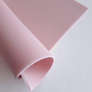 Könnte beinhalten: Eine Rolle hellrosa Stoff mit weißem Rand ist teilweise abgerollt und zeigt ein flaches Blatt aus dem gleichen Material. Der Stoff hat eine glatte Textur und eine weiche, gedämpfte Farbe, geeignet für verschiedene Bastelprojekte.