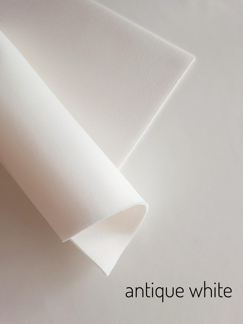 Puede incluir: Primer plano de dos hojas de papel blanco antiguo, una enrollada y la otra plana. El papel tiene un aspecto suave y texturizado. Las palabras "antique white" est&aacute;n impresas en negro, con una fuente sans-serif, en la esquina inferior derecha de la imagen.