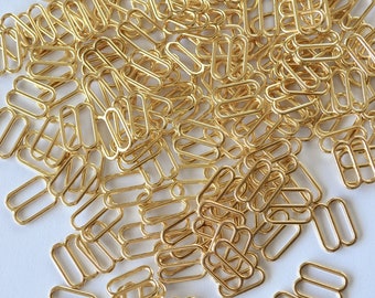 May include: A large pile of gold-coloured metal slider buckles. The buckles are oval-shaped with two loops.