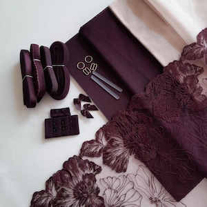 Könnte beinhalten: Eine Auswahl an Materialien zur Herstellung von Dessous, darunter bordeauxfarbener Stoff, Spitzenbesatz mit floralen Designs, Gummiband und BH-Verschlüsse. Ebenfalls enthalten sind kleine goldfarbene Ringe und Schieber sowie winzige bordeauxfarbene Schleifen. Die Artikel sind auf einer weißen Oberfläche angeordnet.