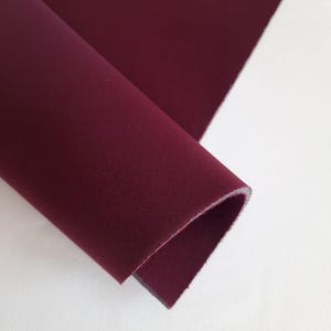 May include: A close-up of a rolled-up piece of burgundy fabric. The fabric is a solid color and appears to be a medium-weight material. The edges of the fabric are visible, showing a layered construction. The background is a plain white surface.