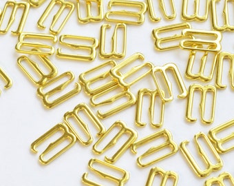 May include: A selection of gold-coloured bra strap hooks, scattered on a white surface. The hooks are made of metal and have a rectangular shape with a curved hook on each end. These are used for lingerie and sewing projects.