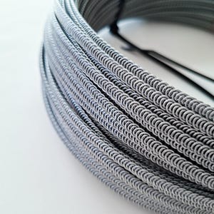 Può includere: Un fascio arrotolato di filo metallico grigio, avvolto strettamente e fissato con una fascia nera. Il filo ha un aspetto strutturato, simile a una catena, con un motivo ripetuto di piccoli anelli collegati. Il colore generale è un grigio tenue, con riflessi che creano un senso di profondità.