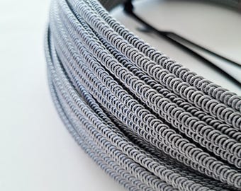 May include: A coiled bundle of grey metal wire, tightly wound and secured with a black band. The wire has a textured, chain-like appearance, with a repeating pattern of small, connected loops. The overall colour is a muted grey, with highlights creating a sense of depth.
