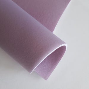May include: A close-up of a rolled-up piece of fabric in a muted lavender color. The fabric has a smooth texture on the top surface and a white underside. The fabric is on a white surface.