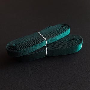 May include: Two spools of dark green ribbon, bound together with white string. The ribbon is tightly wound, showing a textured surface. The background is a solid black.