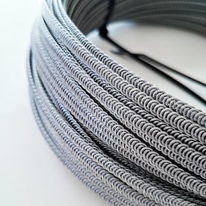 Może przedstawiać: Zbliżenie na zwinięty w krąg szary drut metalowy, mocno owinięty i zabezpieczony czarną taśmą. Drut ma teksturowany, pętelkowy wzór, tworzący powtarzający się wzór półokręgów. Obraz szczegółowo ukazuje detale metalowego drutu.