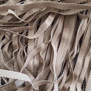 May include: Close-up of a pile of beige elastic trim with a scalloped edge and a white stitched detail. The trim is soft and flexible, ideal for sewing and crafting projects.