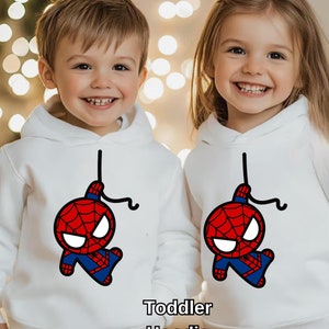 Puede incluir: Dos ni&ntilde;os con sudaderas blancas con una imagen de dibujos animados de Spiderman colgando boca abajo. Las sudaderas dicen "Toddler Hoodie" en la parte inferior.