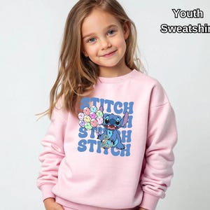 May include: Pink youth sweatshirt featuring a blue Stitch character holding colorful balloons with the word "Stitch" repeated in a retro font.