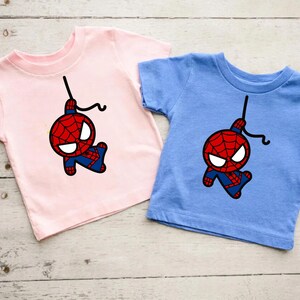 Puede incluir: Dos camisetas con una imagen de dibujos animados de Spiderman colgando boca abajo en una telara&ntilde;a. La camiseta de la izquierda es rosa y la camiseta de la derecha es azul.