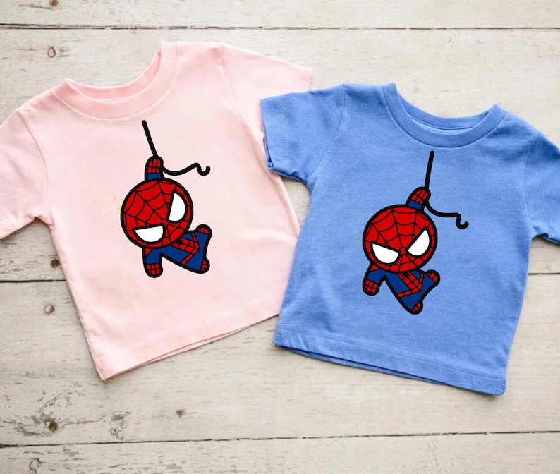 Puede incluir: Dos camisetas con una imagen de dibujos animados de Spiderman colgando boca abajo en una telara&ntilde;a. La camiseta de la izquierda es rosa y la camiseta de la derecha es azul.