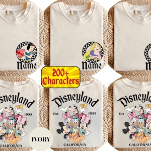 May include: Ivory-colored t-shirts featuring Disney characters and the Disneyland logo. The shirts display Snow White, Rapunzel, and Elsa designs, along with a group of Disney characters. The text "Name" appears below each princess.