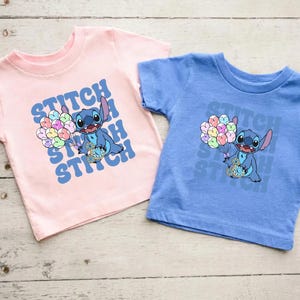 May include: Two t-shirts featuring Stitch from Lilo & Stitch. The pink shirt has a blue Stitch holding balloons with the text "Stitch Stitch Stitch" in a retro font. The blue shirt has a blue Stitch holding balloons with the text "Stitch Stitch Stitch" in a retro font.
