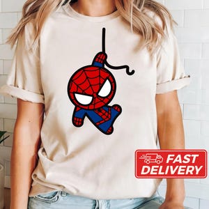 Puede incluir: Una camiseta beige con una imagen de dibujos animados de Spiderman balance&aacute;ndose en una telara&ntilde;a. La imagen es en negro, rojo y azul.