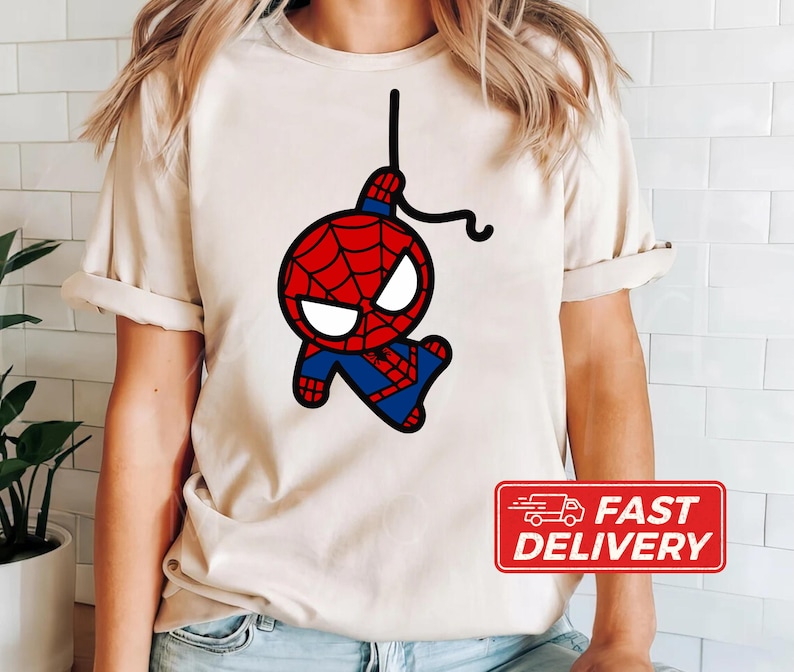 Puede incluir: Una camiseta beige con una imagen de dibujos animados de Spiderman balance&aacute;ndose en una telara&ntilde;a. La imagen es en negro, rojo y azul.