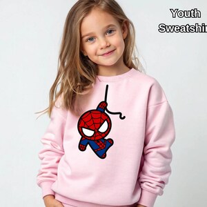 Puede incluir: Sudadera rosa para j&oacute;venes con una imagen de dibujos animados de Spiderman colgando de una telara&ntilde;a. El texto "Youth Sweatshirt" est&aacute; en la esquina superior derecha de la imagen.